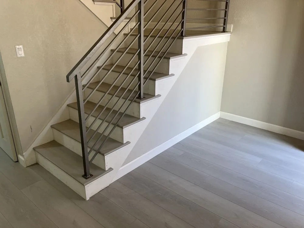 Light gray vinyl plank flooring on stairs with white risers and metal railing by The Flooring Design Center